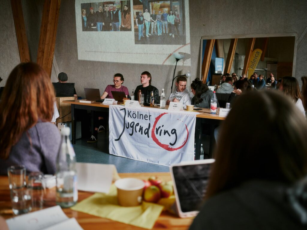 junge Menschen sitzen an einem Tisch, davor das Banner des Kölner Jugendrings